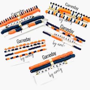 May include: Several bracelets in orange, navy, white, and gold are displayed on white cards. The cards read "Gameday" and "YOUR TEAM" in black script. Some bracelets feature beads, others flat disc beads.