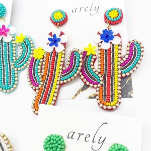 May include: A pair of colorful beaded cactus earrings with a white flower and yellow center. The earrings are red, orange, yellow, green, blue, and black.