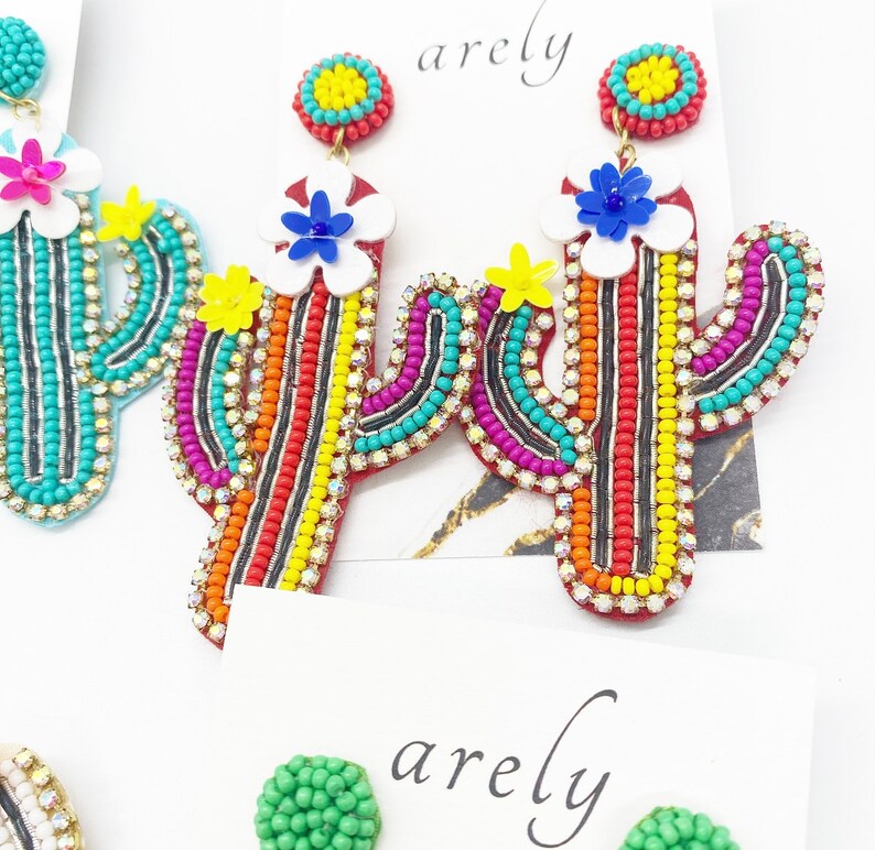 May include: A pair of colorful beaded cactus earrings with a white flower and yellow center. The earrings are red, orange, yellow, green, blue, and black.