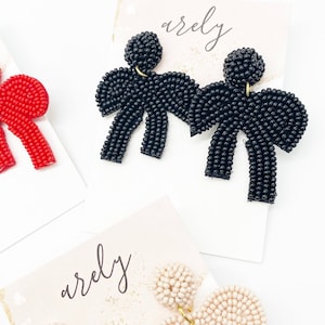 May include: Three pairs of beaded bow earrings in red, black, and cream. The earrings are displayed on white cards with the word "arely" written on them.