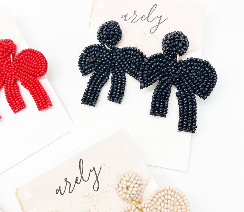 May include: Three pairs of beaded bow earrings in red, black, and cream. The earrings are displayed on white cards with the word "arely" written on them.