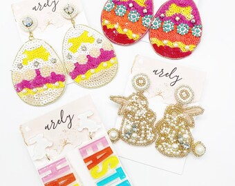 May include: A set of four pairs of Easter-themed earrings. The earrings are made of sequins, beads, and pearls. The earrings are in the shape of eggs, bunnies, and the words "Happy Easter".