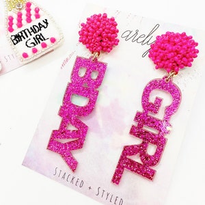May include: Pink glitter earrings with the words "Birthday Girl" on them. The earrings have pink bead accents.