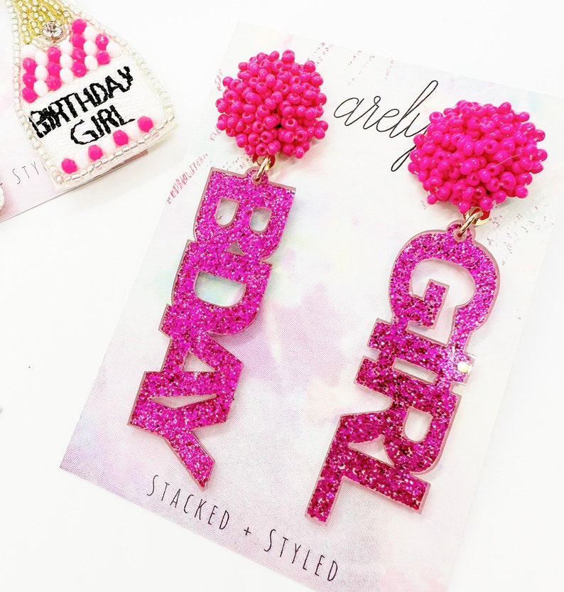 May include: Pink glitter earrings with the words "Birthday Girl" on them. The earrings have pink bead accents.