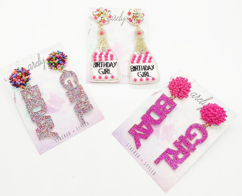 May include: Three pairs of pink and white glitter earrings. The first pair features the word "Birthday Girl" in pink glitter. The second pair features the word "Girl" in pink glitter. The third pair features the word "Ava" in pink glitter.