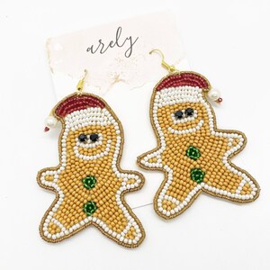 May include: A pair of gingerbread man-shaped earrings with white, gold, red, and green beads. The earrings are made of beads and have a gold hook.