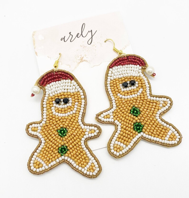 May include: A pair of gingerbread man-shaped earrings with white, gold, red, and green beads. The earrings are made of beads and have a gold hook.