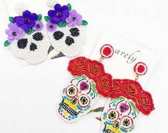 May include: A pair of beaded skull earrings. One pair is white with purple flowers and the other pair is white with red flowers. The earrings are both in the shape of a skull with a beaded design.