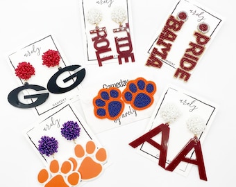 May include: A selection of handmade earrings featuring various college team logos and colours, including paw prints, letters, and words such as "Roll Tide" and "Bama Pride".