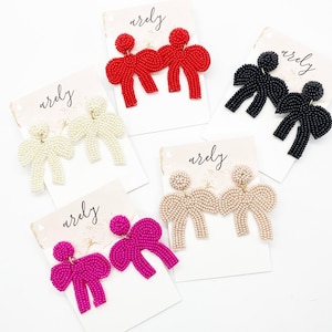 May include: Five pairs of beaded bow earrings in different colors. The earrings are displayed on white cards with the word "arely" written on them. The colors of the earrings are red, black, white, beige, and pink.