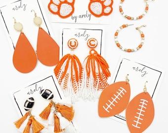 May include: A set of orange and white tassel earrings with a football and paw print charm. The earrings are labelled "Gameday by Arely".