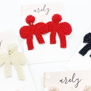 May include: A pair of red beaded bow earrings. The bows are made of small red beads and are connected in the center with a gold-colored metal piece. The earrings are displayed on a white card with the word "arely" written on it.