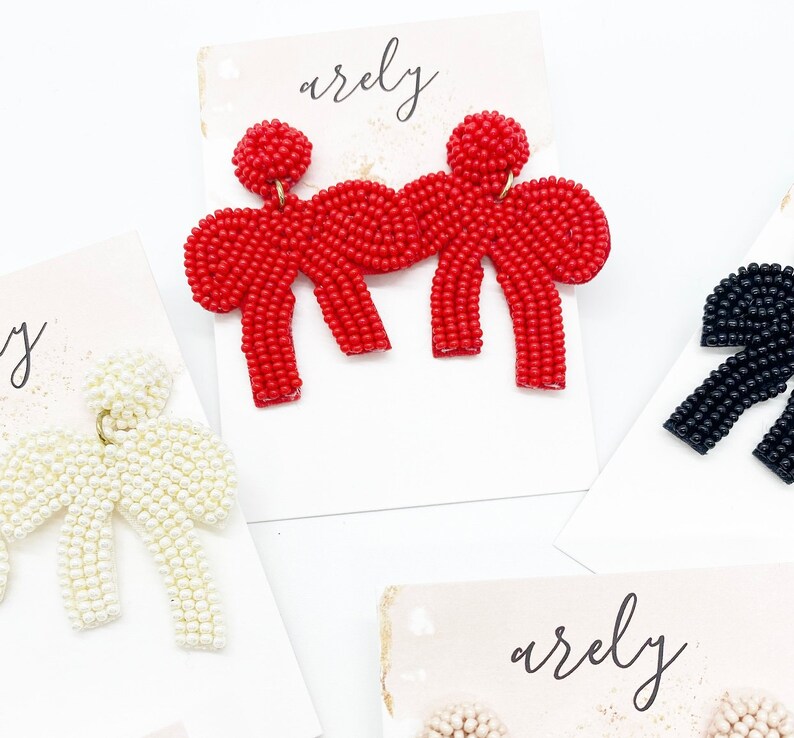 May include: A pair of red beaded bow earrings. The bows are made of small red beads and are connected in the center with a gold-colored metal piece. The earrings are displayed on a white card with the word "arely" written on it.