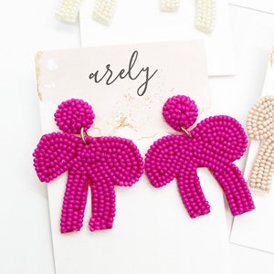 May include: A pair of pink beaded earrings shaped like bows. The earrings are on a white background with the word "arely" written in black.