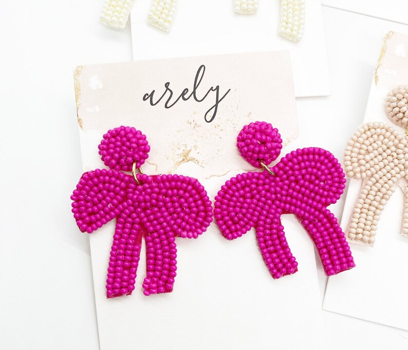 May include: A pair of pink beaded earrings shaped like bows. The earrings are on a white background with the word "arely" written in black.