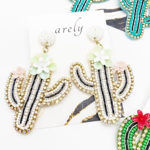 May include: A pair of dangle earrings featuring a cactus design with white and black beads, green and pink flower accents, and gold-tone hardware. The earrings are labeled "arely".