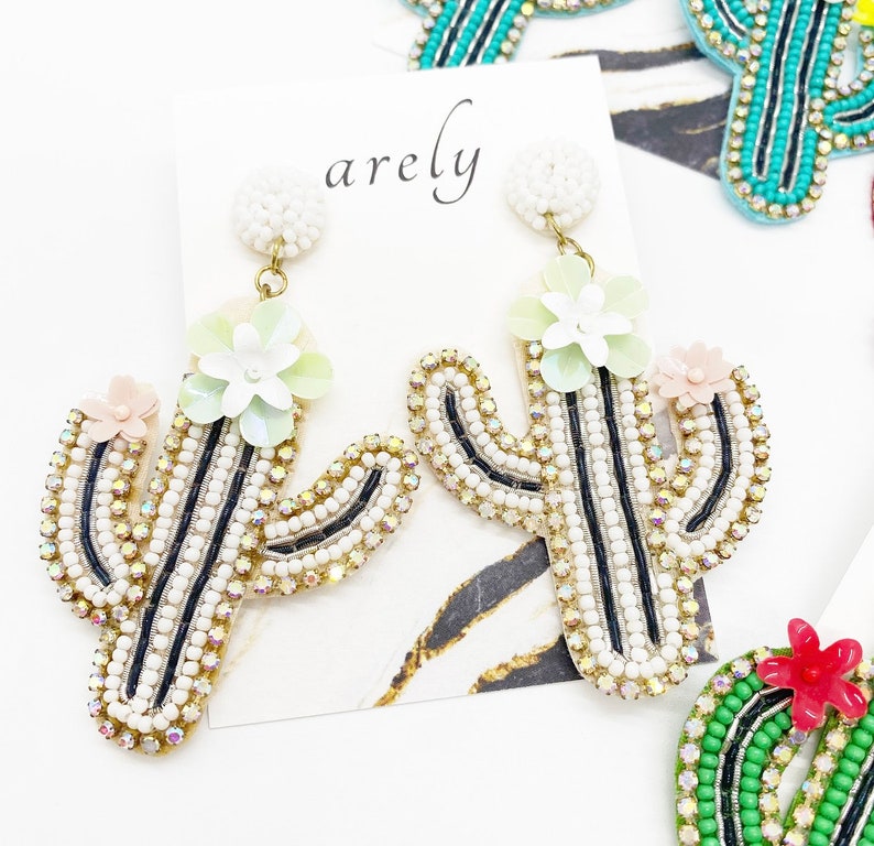 May include: A pair of dangle earrings featuring a cactus design with white and black beads, green and pink flower accents, and gold-tone hardware. The earrings are labeled "arely".