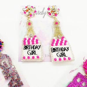 May include: A pair of white beaded earrings shaped like champagne bottles with pink and white beads. The bottles have the text "Birthday Girl" embroidered on them. The earrings have a crystal embellishment at the top.