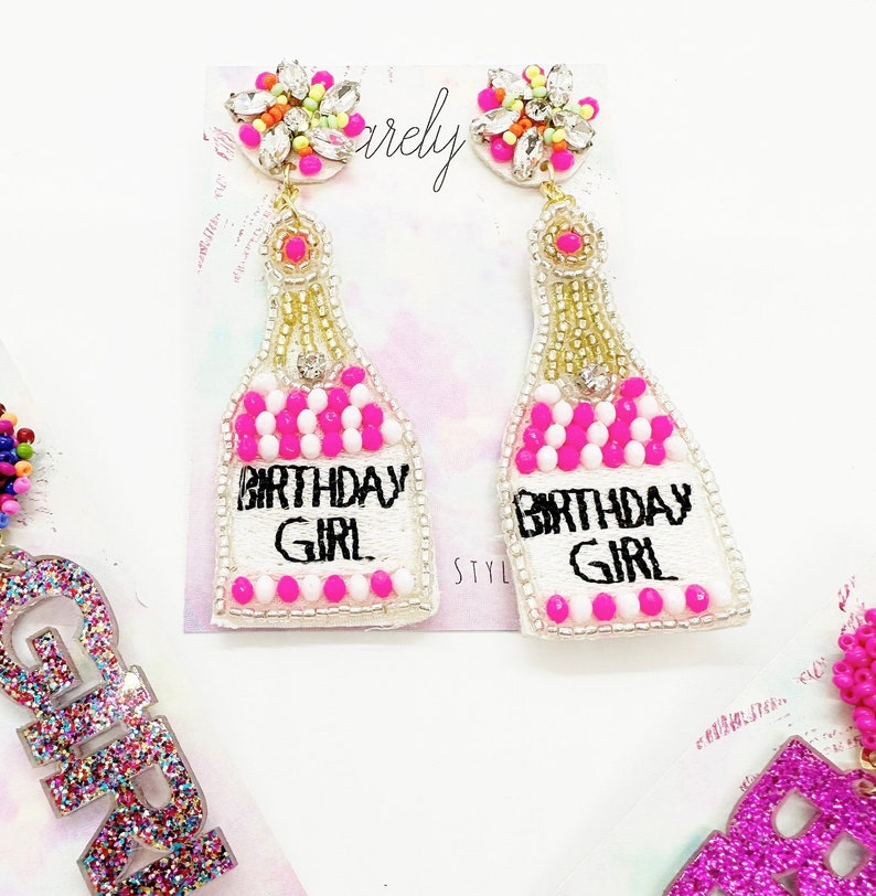 May include: A pair of white beaded earrings shaped like champagne bottles with pink and white beads. The bottles have the text "Birthday Girl" embroidered on them. The earrings have a crystal embellishment at the top.