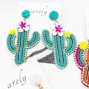 May include: A pair of turquoise cactus earrings with white flowers and yellow accents. The earrings are beaded and have a gold-tone hook.
