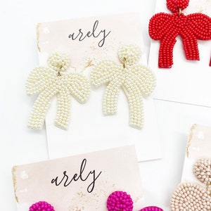 May include: A pair of white pearl beaded bow earrings on a white card with the word "arely" written on it.