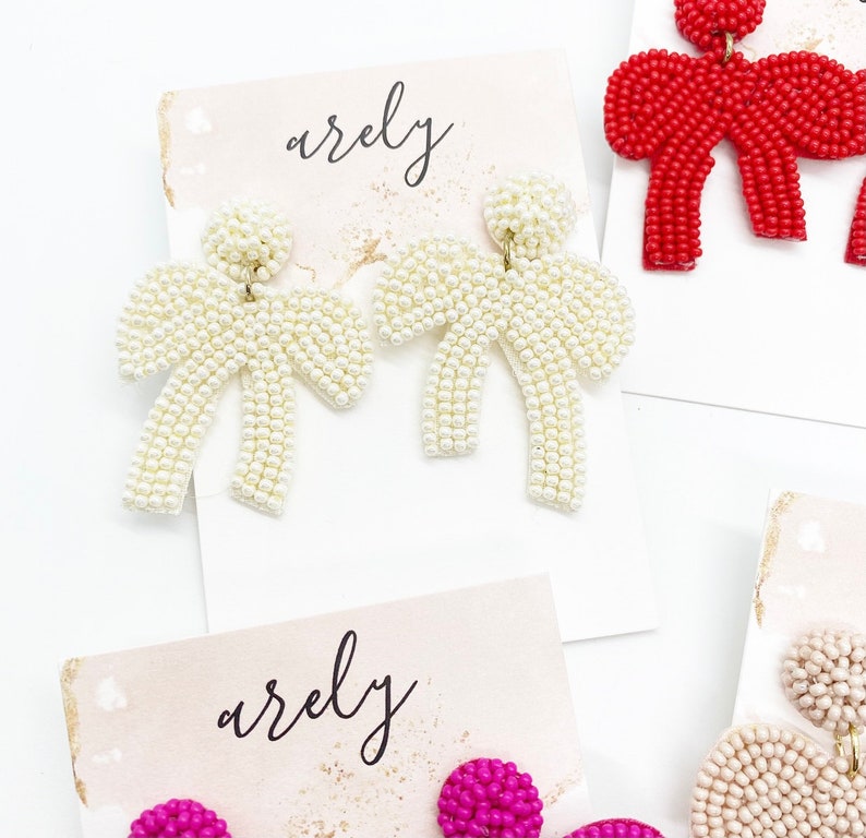 May include: A pair of white pearl beaded bow earrings on a white card with the word "arely" written on it.