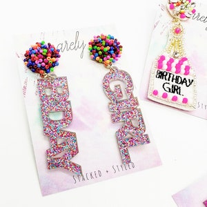May include: A pair of glittery pink earrings with the words "Birthday Girl" and "Birthday" spelled out. Each earring has a colourful beaded ball at the top.