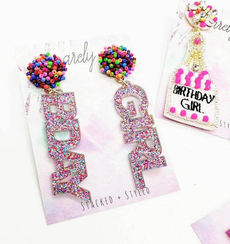May include: A pair of glittery pink earrings with the words "Birthday Girl" and "Birthday" spelled out. Each earring has a colourful beaded ball at the top.