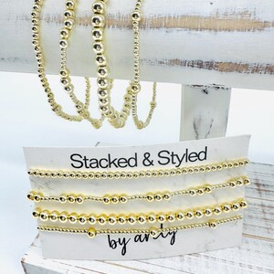 May include: Gold beaded bracelets displayed on a white wooden stand. The bracelets vary in bead size and are presented on a card that reads "Stacked & Styled by arvy". The jewelry is a warm gold color.