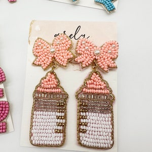 May include: A pair of pink beaded earrings with a bow design and a gold-toned bottle charm. The bottle charm is also beaded and has a white and pink design.