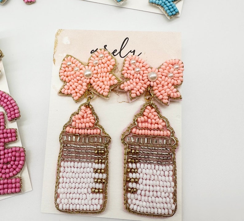 May include: A pair of pink beaded earrings with a bow design and a gold-toned bottle charm. The bottle charm is also beaded and has a white and pink design.