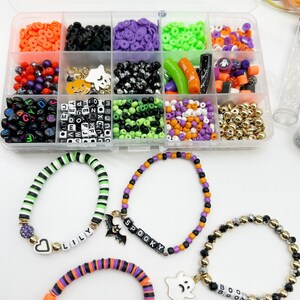 May include: A clear plastic storage box filled with various Halloween-themed beads, charms, and spacers. The box includes black, white, green, orange, and purple beads, as well as letter beads, pumpkin charms, ghost charms, and bat charms. Three bracelets are shown, each with a different charm and name: "Lilly", "Spooky", and "Boo".