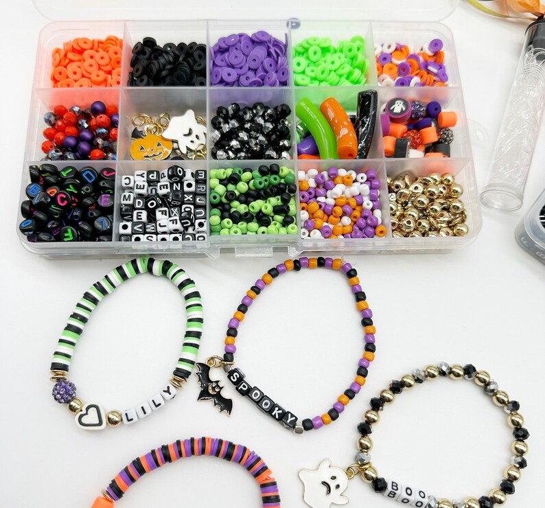 May include: A clear plastic storage box filled with various Halloween-themed beads, charms, and spacers. The box includes black, white, green, orange, and purple beads, as well as letter beads, pumpkin charms, ghost charms, and bat charms. Three bracelets are shown, each with a different charm and name: "Lilly", "Spooky", and "Boo".