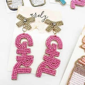 May include: Pink beaded earrings with a gold bow and a rhinestone in the center. The earrings spell out the word "GIRL" in pink beads.