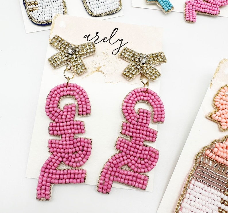 May include: Pink beaded earrings with a gold bow and a rhinestone in the center. The earrings spell out the word "GIRL" in pink beads.