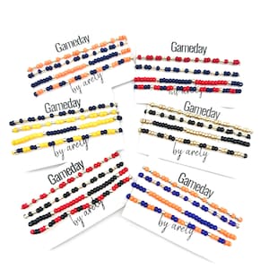 Puede incluir: Varios pasadores para el cabello con cuentas en varias combinaciones de colores, incluyendo naranja, azul marino, rojo, negro, amarillo y dorado. Cada pasador se muestra en una tarjeta blanca con el texto "Gameday" y "by arely".