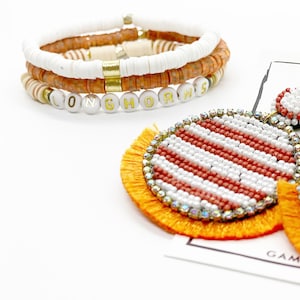 Puede incluir: Un conjunto de pulseras de cuentas naranja y blancas con la palabra "Longhorns" escrita en letras doradas. Las pulseras se combinan con un par de pendientes de cuentas rayadas naranja y blancas con borlas naranjas.