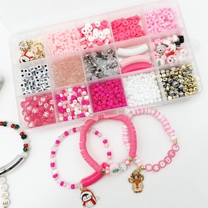 May include: A clear plastic storage box filled with various pink, white, and gold beads, charms, and letters. Three bracelets are shown with pink and white beads, a pink charm, and a gold reindeer charm. The bracelets spell out "Maddie" and "Berry".