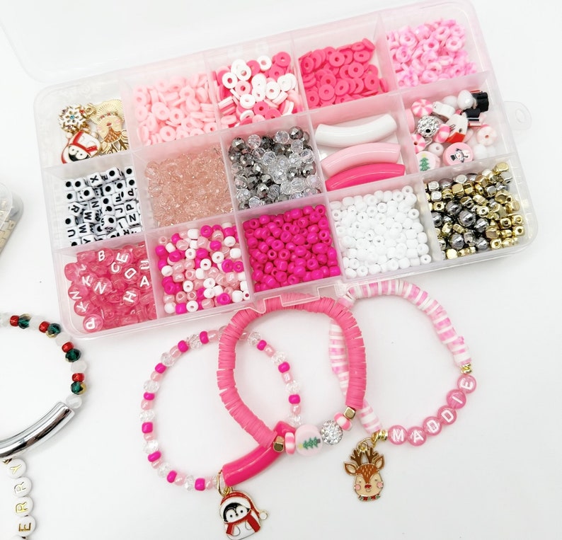 May include: A clear plastic storage box filled with various pink, white, and gold beads, charms, and letters. Three bracelets are shown with pink and white beads, a pink charm, and a gold reindeer charm. The bracelets spell out "Maddie" and "Berry".
