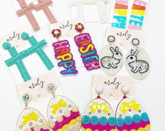 May include: A selection of colourful beaded earrings featuring various designs, including crosses, Easter eggs, bunnies, and the words "Happy" and "Easter".