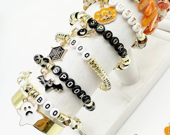 Könnte beinhalten: Fünf Halloween-Armbänder mit goldenen Perlen und schwarzen Buchstabenperlen, die "BOO", "SPOOKY" und "CUSTOM" buchstabieren. Die Armbänder haben Charms, darunter eine Spinne, eine Fledermaus, ein Geist und ein Kürbis.