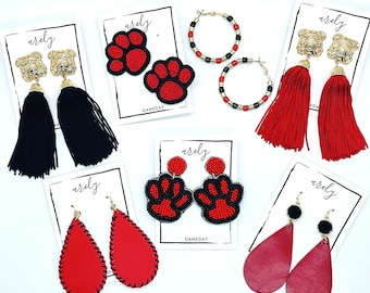 Peut inclure: Assortiment de boucles d'oreilles en perles rouges et noires avec différents motifs, dont des empreintes de pattes, des bouledogues et des gouttes de glands. Les boucles d'oreilles sont présentées sur des cartes blanches avec le mot "ardly" et "GAMEDAY" imprimé dessus.