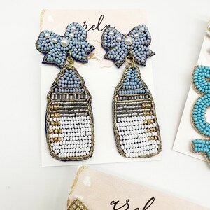 May include: A pair of blue and white beaded earrings with a baby bottle charm and a blue bow with a pearl accent.