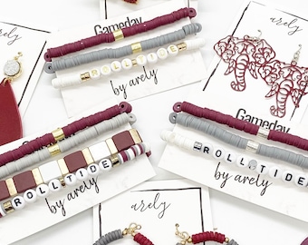 Puede incluir: Tres juegos de pulseras con las palabras "Roll Tide" deletreadas en cuentas blancas. Las pulseras están hechas de cuentas granate, grises y blancas. Las pulseras están en tarjetas blancas con las palabras "Gameday" y "by arely" impresas en ellas.