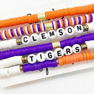 May include: Six colorful beaded bracelets with the words "CLEMSON" and "TIGERS" spelled out in black letters on white beads. The bracelets are made with orange, purple, and white beads.