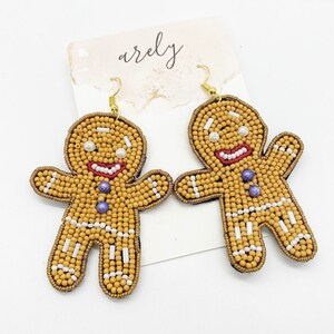 May include: A pair of gingerbread man earrings made of brown, white, and purple beads. The earrings are hanging from gold hooks.