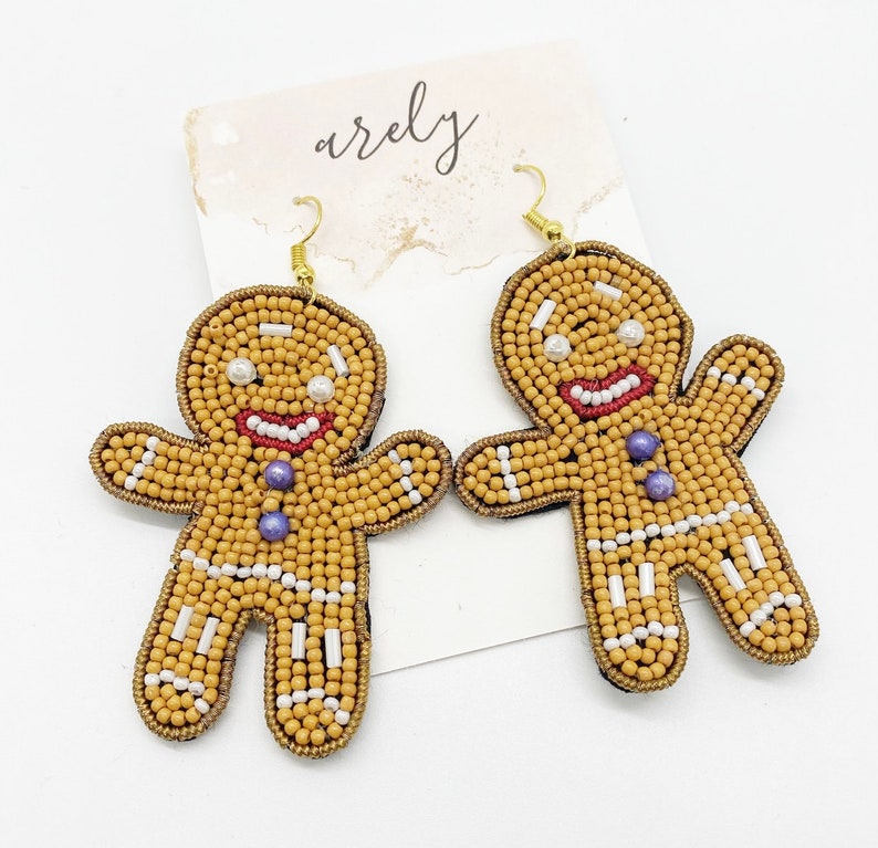 May include: A pair of gingerbread man earrings made of brown, white, and purple beads. The earrings are hanging from gold hooks.