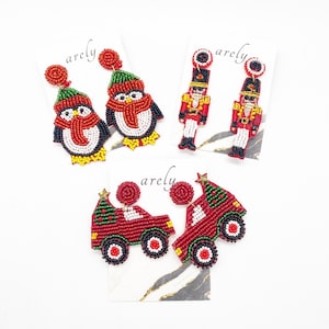 Puede incluir: Tres pares de pendientes de cuentas con temática navideña. El primer par presenta un pingouin rojo y blanco con un sombrero verde. El segundo par presenta un cascanueces rojo y dorado. El tercer par presenta un camión rojo con un árbol de Navidad verde en la parte superior.