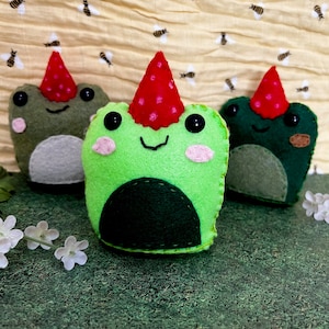 May include: Three felt frog plushies wearing party hats. The frogs are green with black eyes and red mouths. The hats are red with white polka dots.