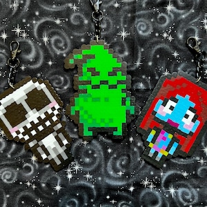 May include: Three pixel art keychains featuring a green monster, a red-haired girl with blue eyes, and a black and white skeleton. The keychains are made of plastic beads and are attached to black metal key rings.